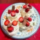 Four mini pavlovas on a plate with fresh cream, strawberries and flakes.